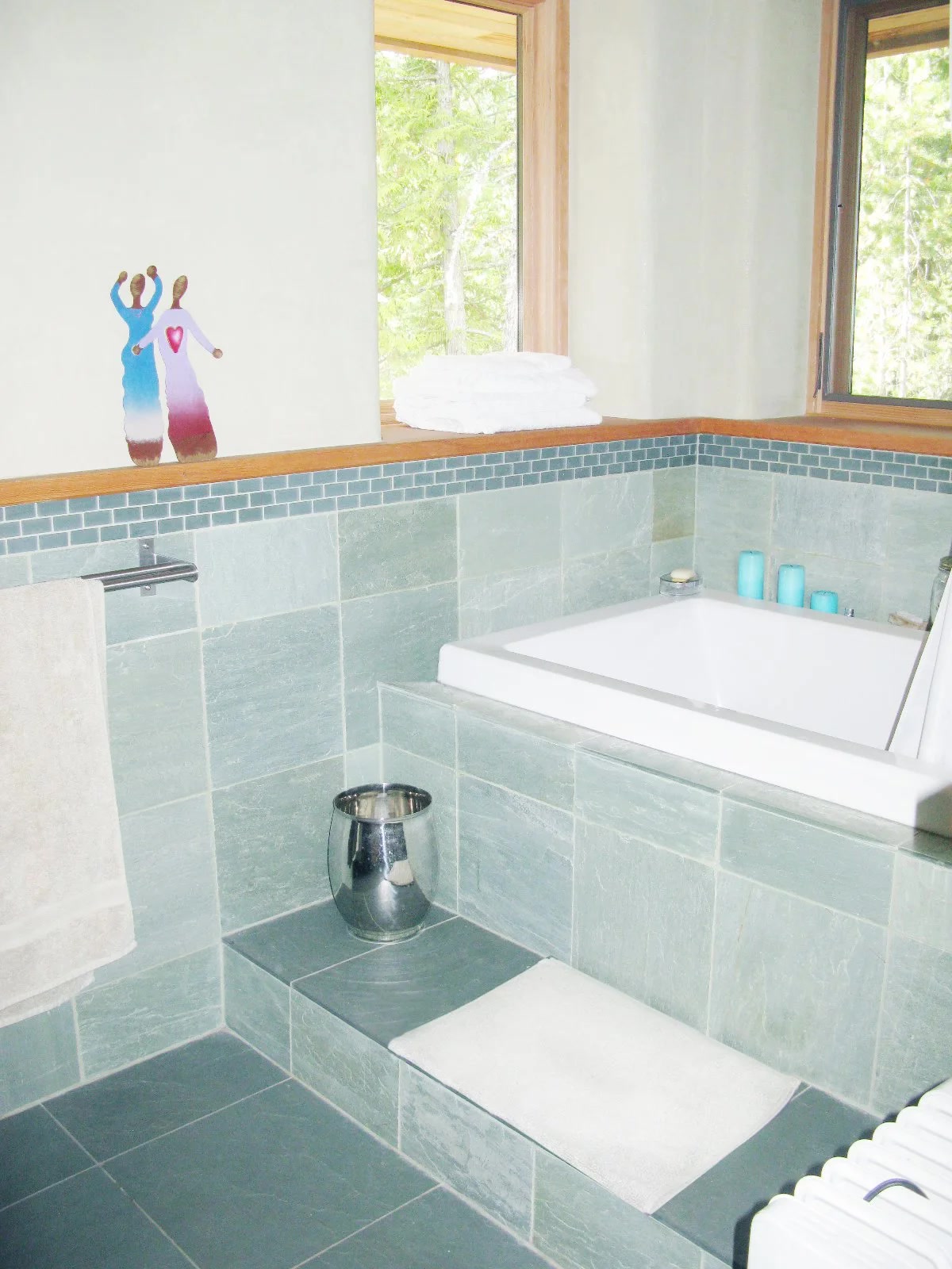 Spa-like bathroom with green slate tile, soaking tub, forest view through window, and natural plaster walls