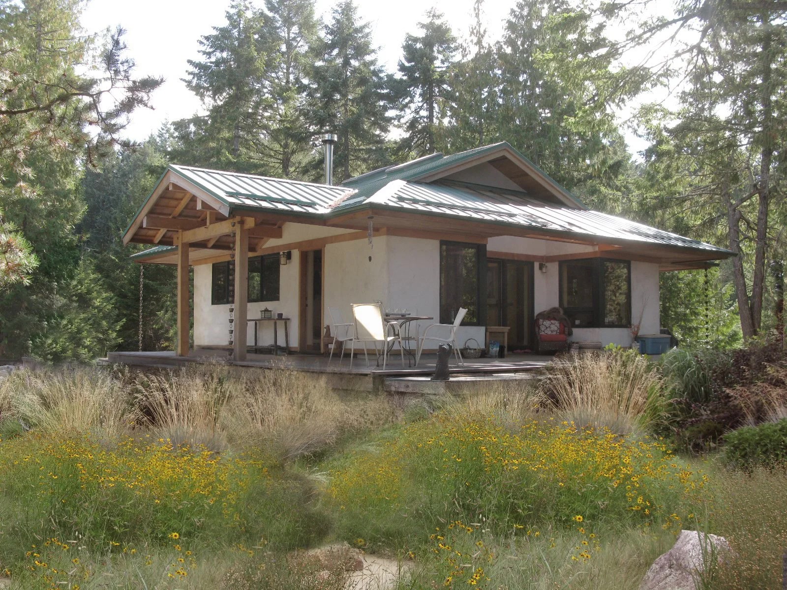 Small natural home with metal roof, timber porch, and natural plaster siding, nestled in evergreen forest with wildflower meadow in foreground